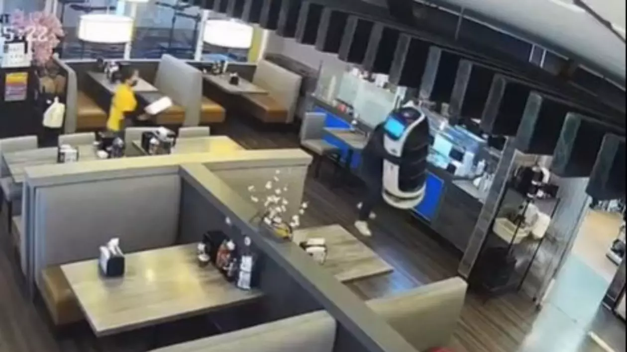 un bărbat cu șapcă, care îi acoperă fața, care cara un chelner robot, în timp ce o angajată îmbracată într-o cămașă galbenă fuge după el