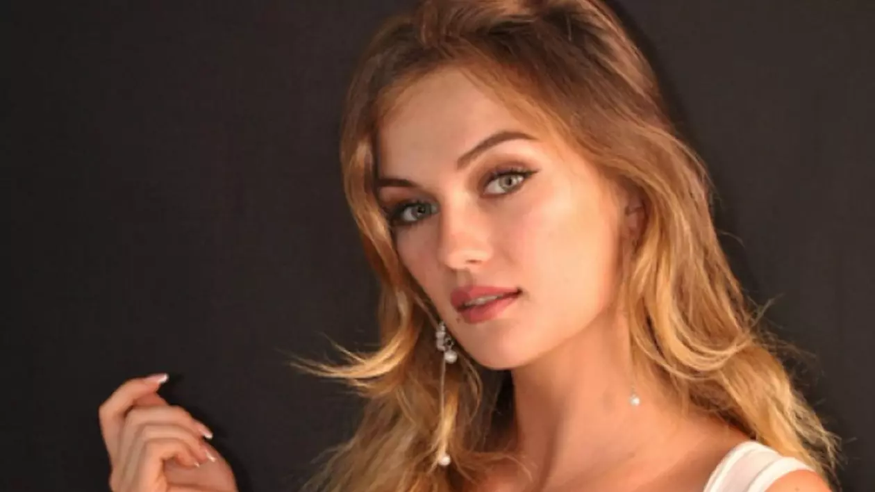 Alexandra, o tânără de 23 de ani, blondă și îmbrăcată cu o bluză albă, pe un fundal gri închis