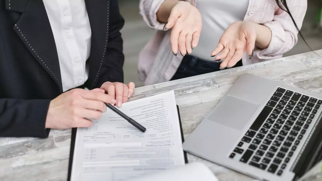 Două femei sunt la o masă pe care este un laptop și un document, stau de vorbă și arată cu pixul pe hârtie, la un interviu de angajare pentru Primăria Timișoara
