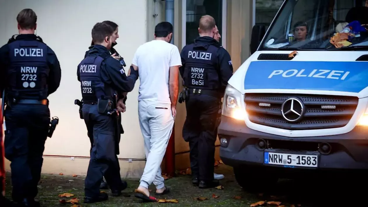 Un bărbat îmbrăcat în alb, cu spatele, este dus de poliția germană în sediul instituției, iar lângă ei se află autospeciala