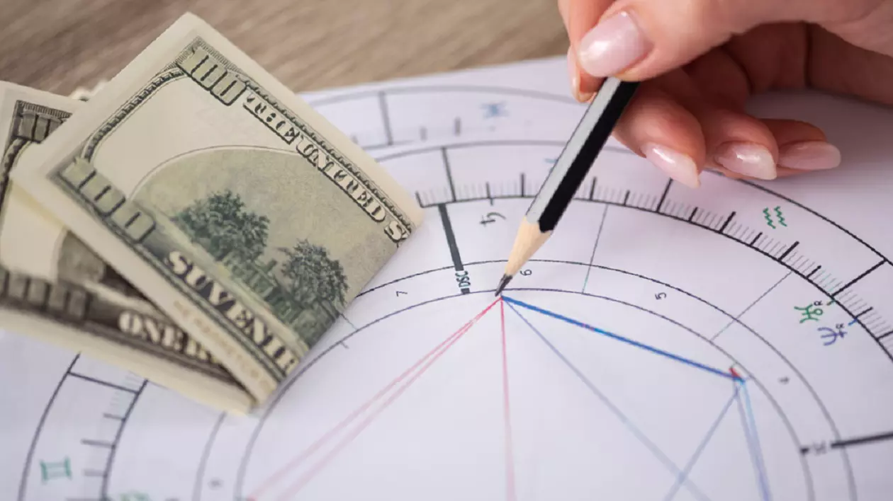 Zodii care au noroc la bani în primăvara lui 2025 - Imagine cu o astrogramaă aşezată pe masă şi peste care se află bancnote de 100 de dolari.