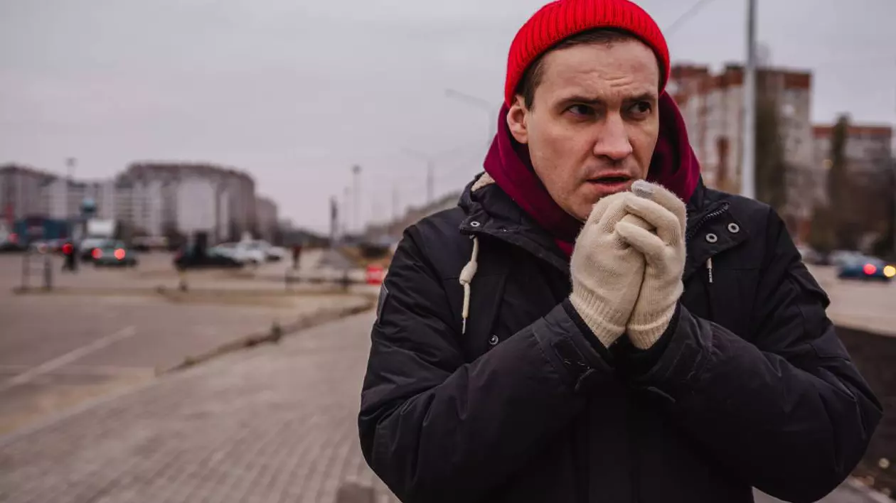 barbat imbracat cu geaca si fes pe cap, manusi. in timp ce isi sufla in pumni de frig