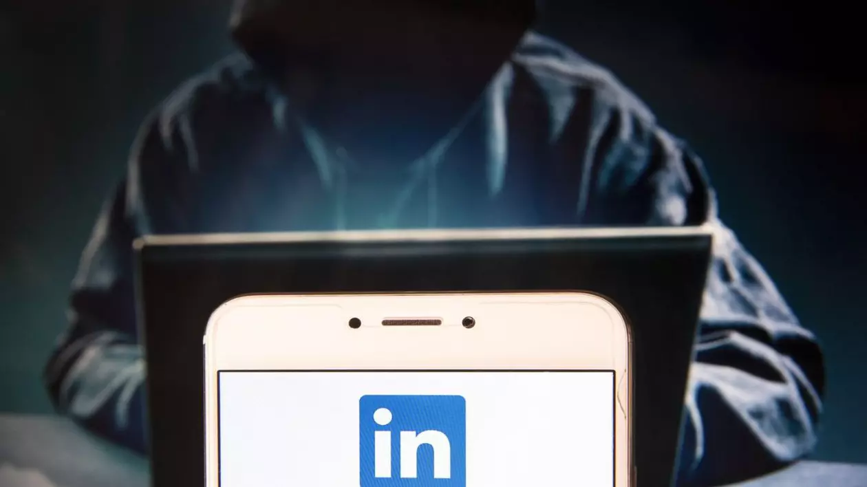hacker îmbrăcat într-un hanorac negru, cu gluga care îi acoperă fața, alături de ecranul unui telefon mobil unui este afișat logo-ul aplicației LinkedIn