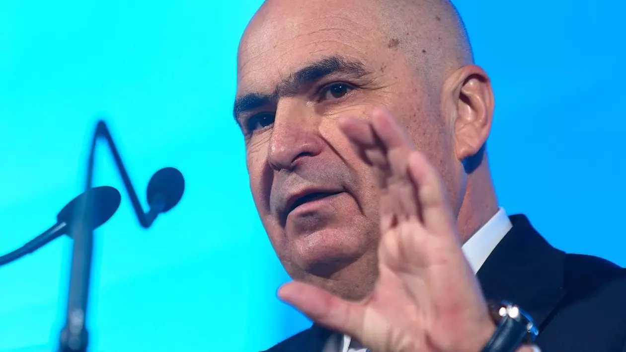 Președintele interimar al României, Ilie Bolojan, surprins când vorbește la microfon și arată cu mâna, pe un fundal bleu-albastru în spate