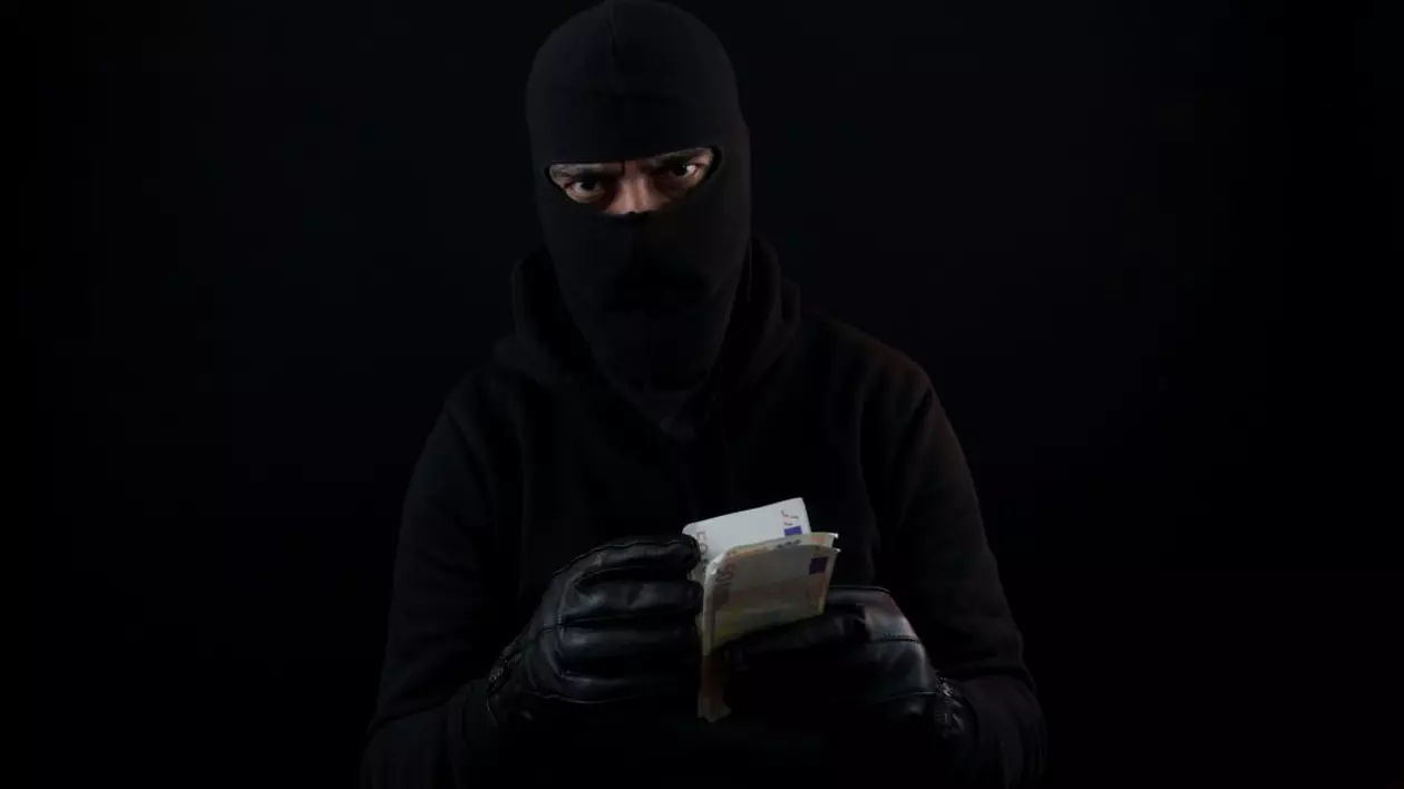 Un bărbat îmbrăcat în negru, cu mănuși și cagulă, ține în mână mai multe banconote de euro și le numără