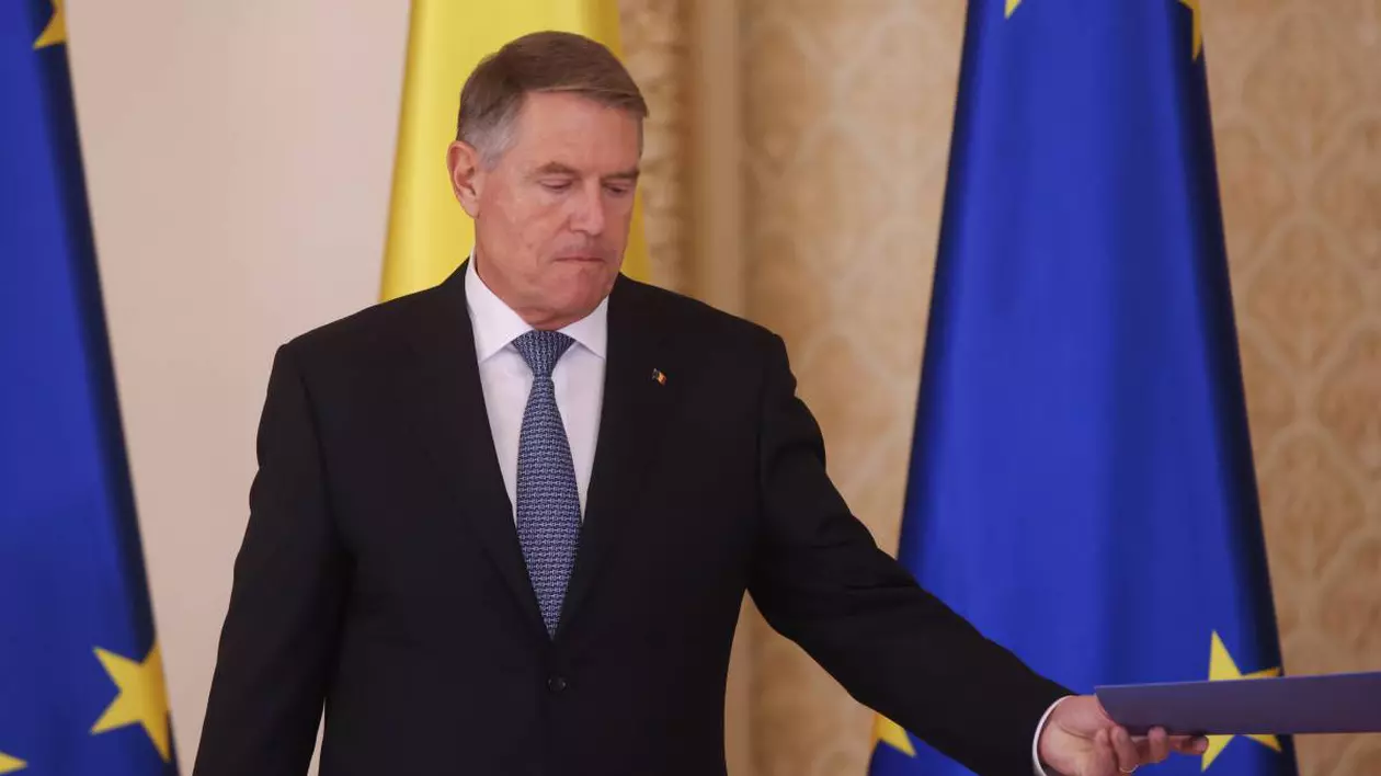 presedintele klaus iohannis cu un document in mana in spatele sau steagul romaniei