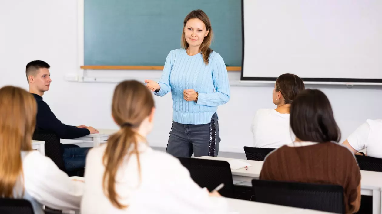 Mai mulți elevi stau în bănci, într-o sală de clasă, iar în fața lor este profesoara, lângă tablă