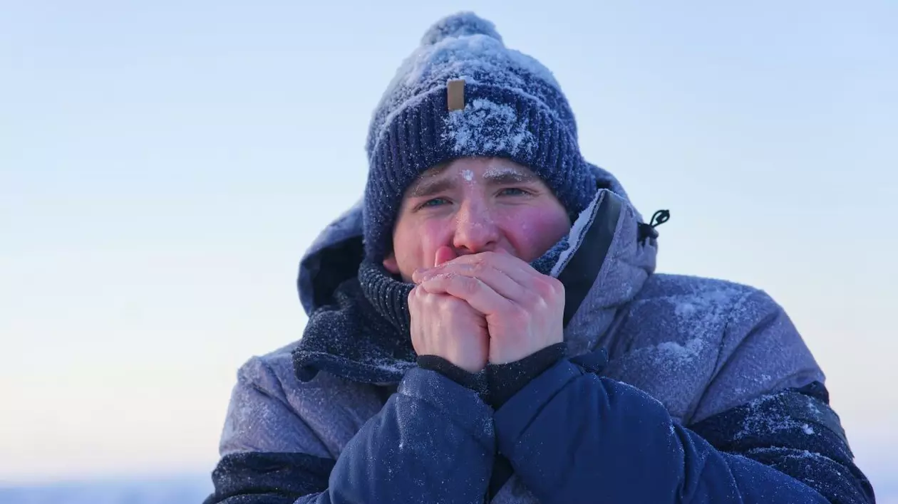 Un bărbat îmbrăcat gros, cu geacă și căciulă, pe care se vede zăpadă, își suflă în mâini din cauza frigului