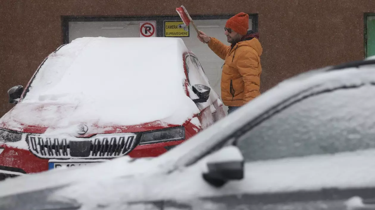 Un bărbat cu geacă și căciulă roșie are în mână o perie cu care vrea să dea jos zăpada de pe o mașină roșie, iar în fața lui se vede un alt autoturism