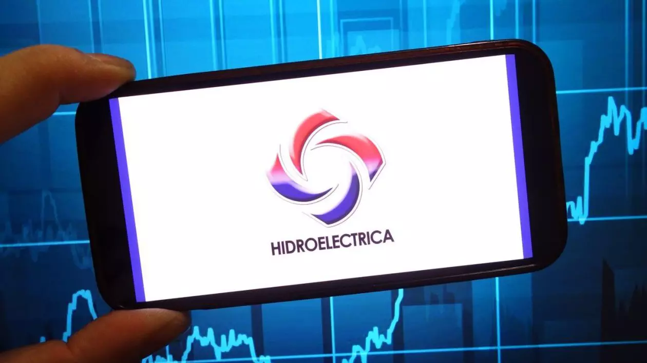 Un om ține în mână un telefon, pe ecranul căruia se vede sigla și numele Hidroelectrica, pe un fundal al cotațiilor bursiere