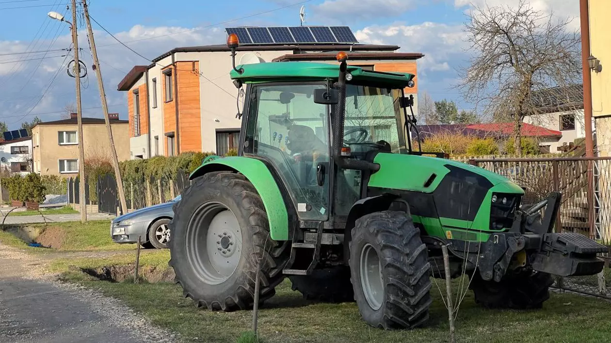 Un tractor verde este parcat pe spațiu verde, în fața unei clădiri, într-o zonă cu blocuri mici și case