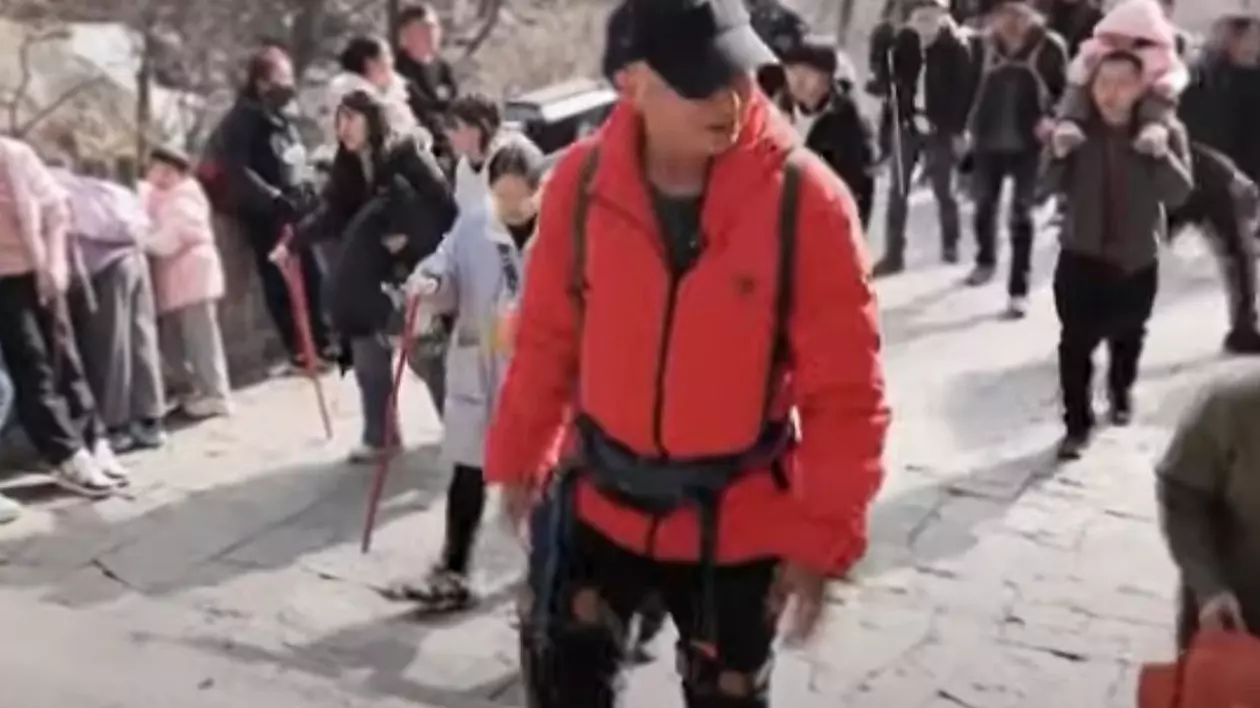 Turist chinez care urcă muntele Tai cu ajutorul exoscheletelor robotizate. Este îmbrăcat de drumeție și are șapcă pe cap