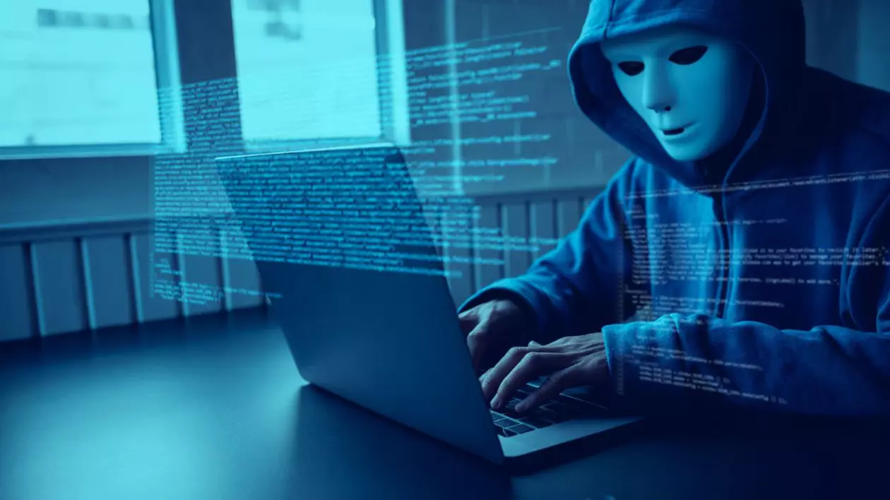 bărbat cu glugă și cu o mască albă pe față, care stă pe laptop. imaginea este acoperită de un text tip cod informatic și este colorată în lumină albastră