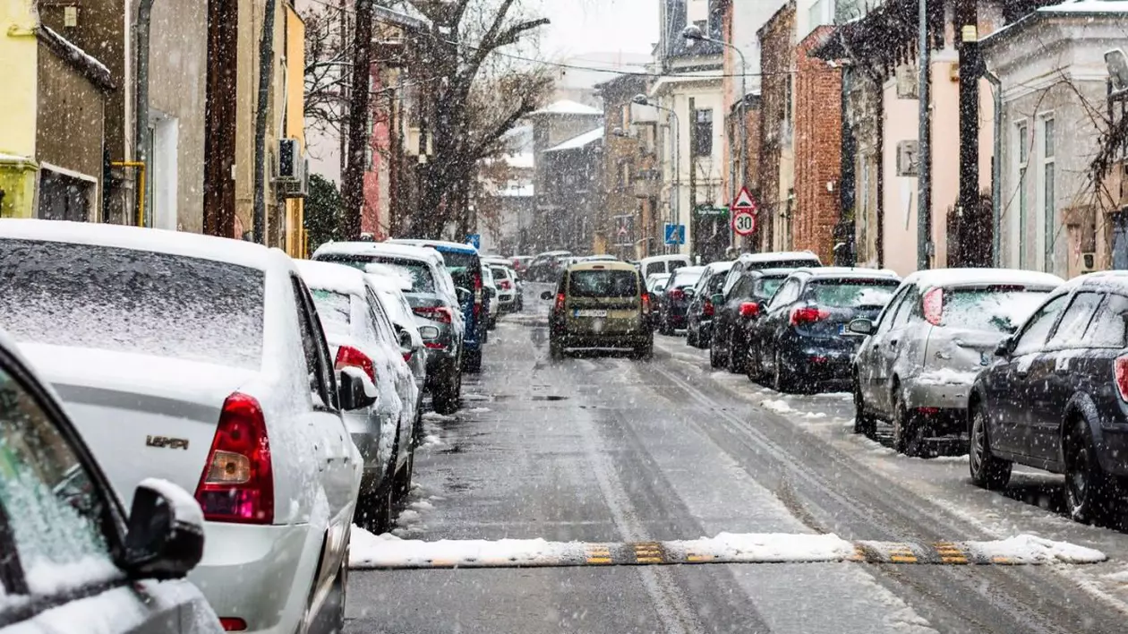 O mașină merge pe o stradă din București într-o zi în care ninge, iar pe trotuarele din stânga și dreapta sunt parcate mașini și se văd case și copaci