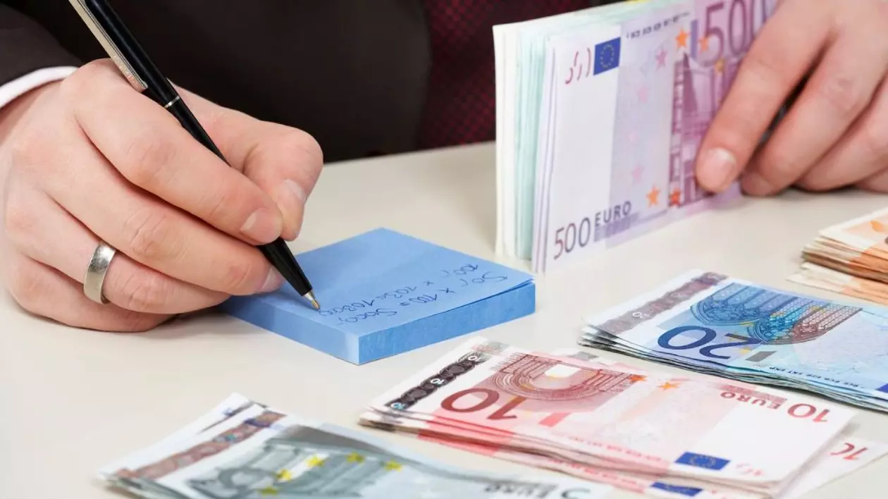 o mână de bărbat care ține un pix face calcule de economoo-cheltuieli si numără banii care sunt alături, pe masă