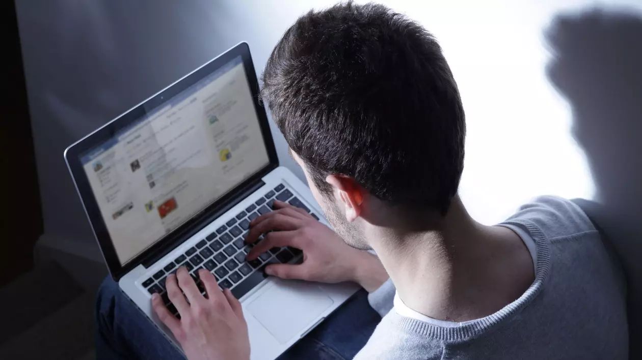 Bărbat surprins în timp ce ține pe picioare un laptop, pe ecranul căruia este afișată platforma Facebook