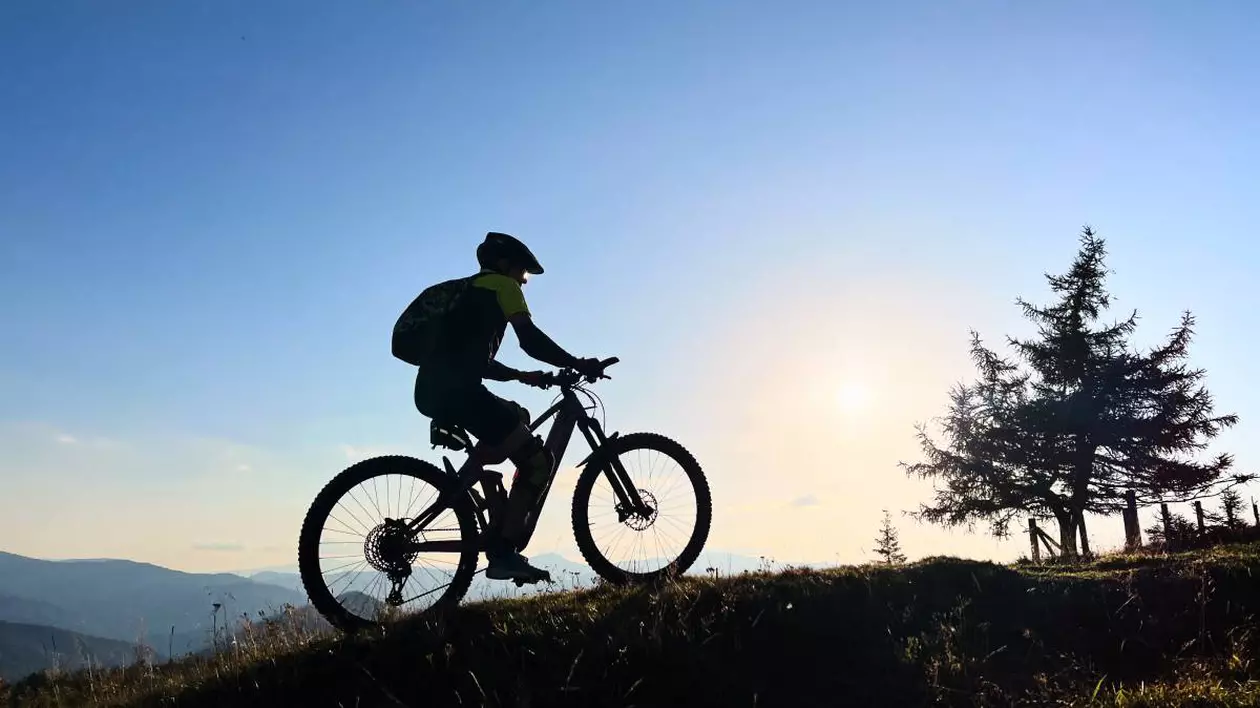 barbat pe o bicicleta pe o poteca montana. in dreapta se observa un brad, fotografia este facuta la apus