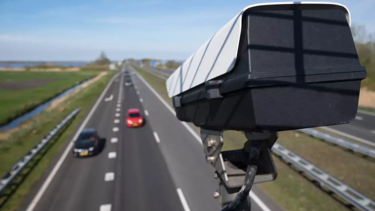 camera video care inregistreaza viteza din trafic. se observa o autostrada cu trei benzi pe sens pe care circula doua masini, una rosie si una neagra