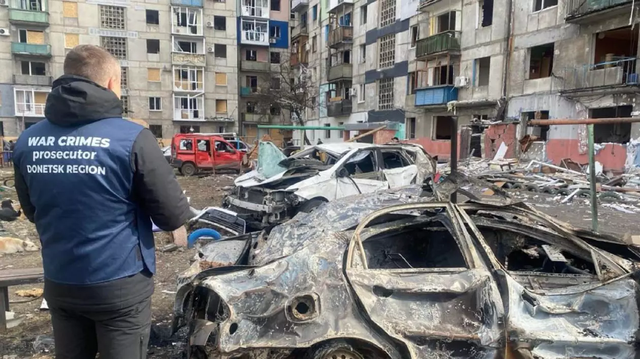 Un procuror care anchetează crime de război, îmbrăcat cu o vestă de culoare albastră, lângă o mașină aflată în parcarea unui bloc, care a explodat în regiunea Donețk din Ucraina