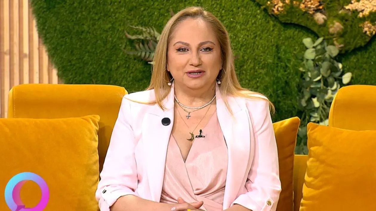 Astrologul Cristina Demetrescu, îmbrăcată cu un sacou alb, stă pe o canapea portocalie în studioul unei emisiuni TV, având în spate un fundal verde