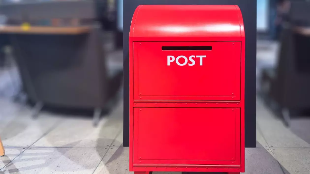 O cutie poștală de culoare roșu aprins este pusă pe o podea cu gresie, într-o clădire. Cutia poștală are un vârf rotunjit, o etichetă mare „POST” și mai multe compartimente