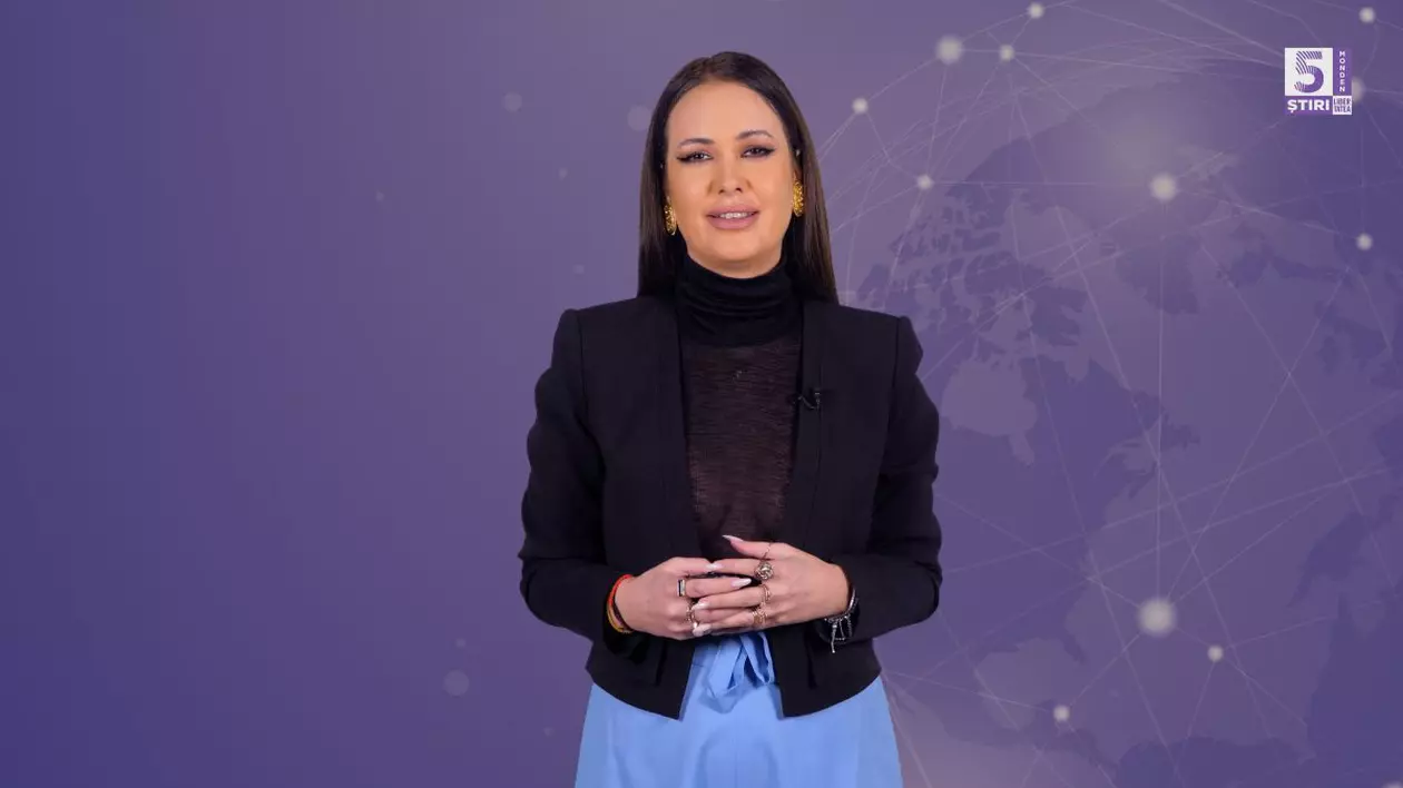 Denisa Macovei, gazda emisiunii 5 știri by Libertatea - Monden, îmbrăcată cu un sacou negru, o bluză neagră și pantaloni de culoare albastră, pe fundal mov
