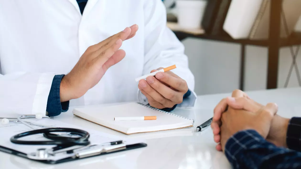 Un doctor îmbrăcat într-un halat alb care ține o țigară în mână, la birou, în fața unui pacient. Pe masă este un stetoscop, un carnet și un pix.