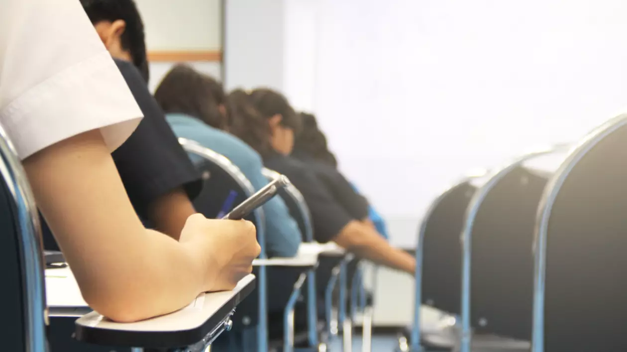 O sală de clasă în care elevii susțin un examen. În prim plan este un elev cu spatele, care are un pix în mână și scrie pe o foaie
