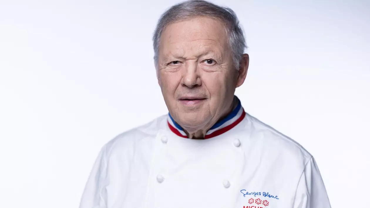 Georges Blanc, în vârstă de 82 de ani, bucătar șef. Poartă un un costum alb de bucătar pe care se vîăd brodate trei stele Mischelin