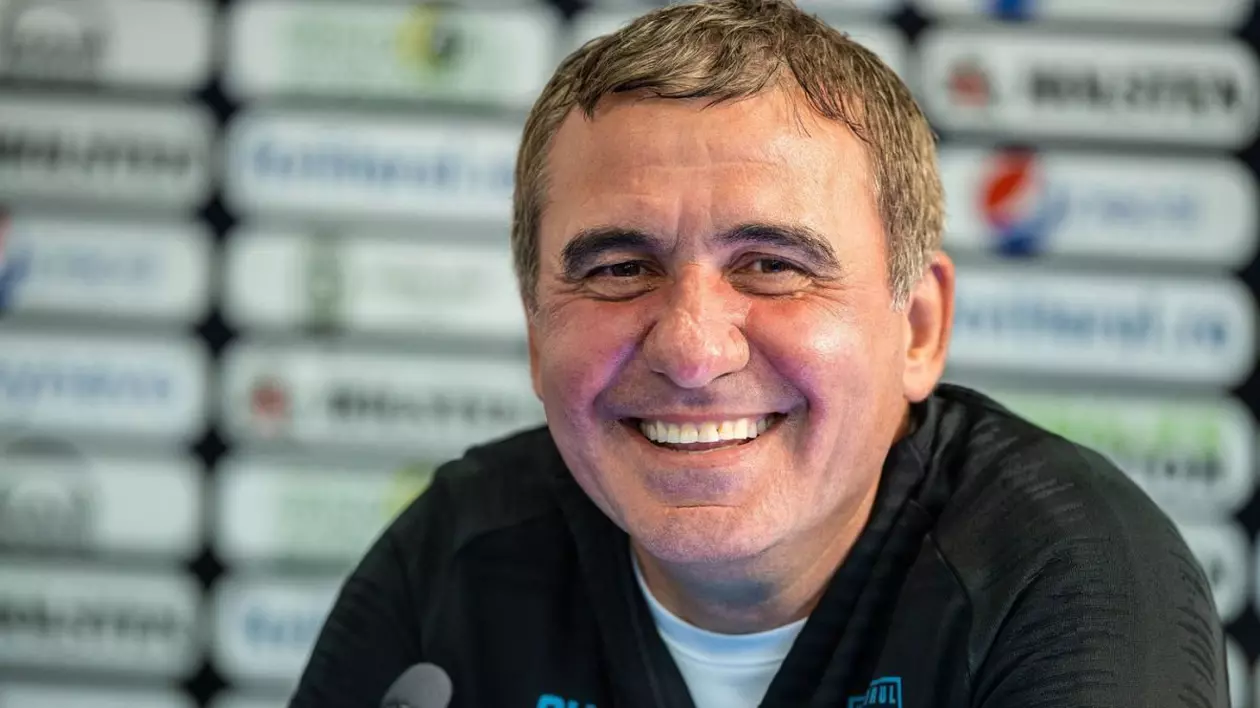 Gheorghe Hagi, îmbrăcat cu o bluză de trening neagră și tricou alb, râde, iar în fața lui este un microfon