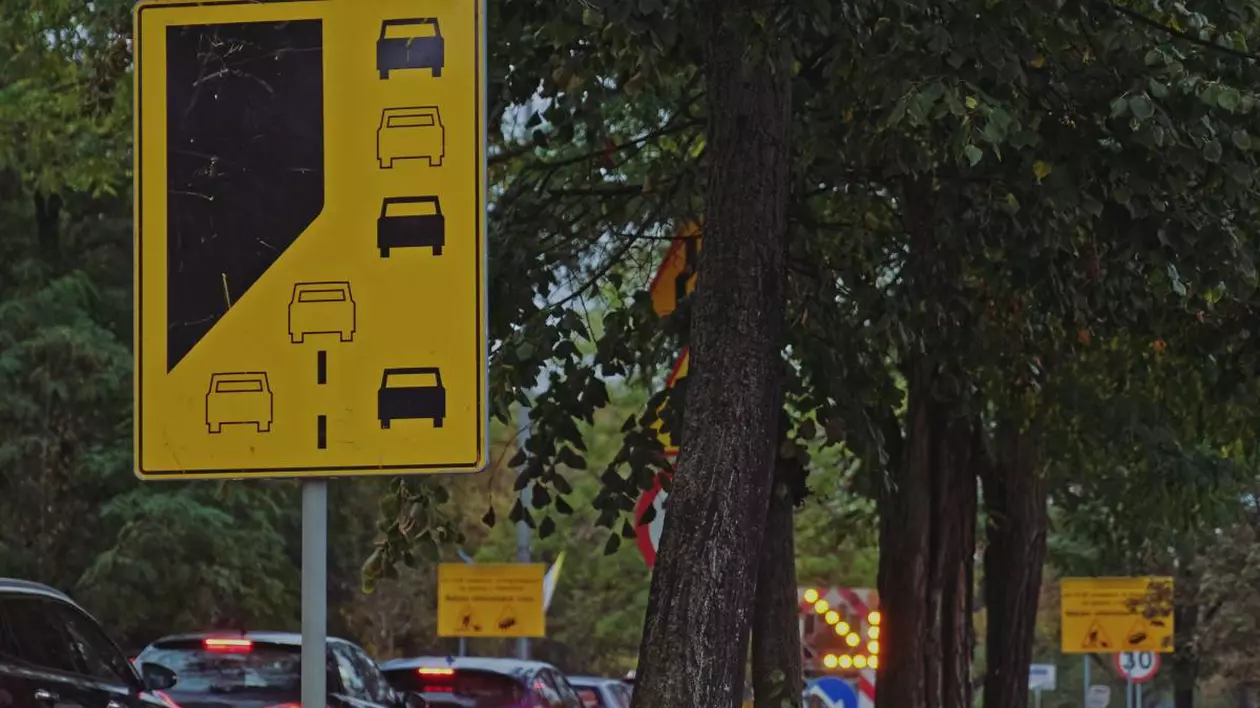 indicator rutier galben care indica regula fermoarului in trafic, pe fundal se observa mai multe masini pe o strada aglomerata din oras. in dreapta sunt niste pomi inalti