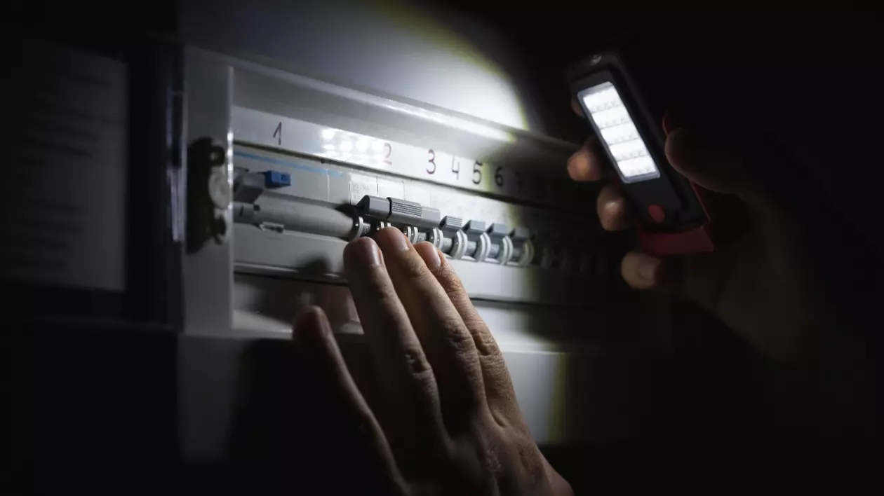Imaginea arată o persoană care verifică sau operează un panou electric într-un mediu întunecat. Persoana folosește o lanternă portabilă cu LED pentru a ilumina siguranțele și întrerupătoarele din panou.