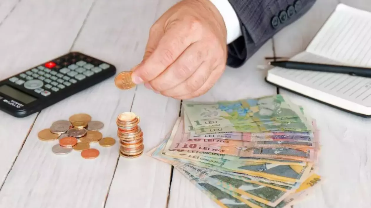 Mâna unui bărbat care pune o monedă pe o masă unde sunt mai mulți bani, o agendă, un pix și un calculator