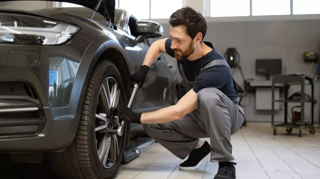 Un mecanic auto îmbrăcat cu salopetă gri și tricou negru, în timp ce strânge prezoanele roții unei mașini negre
