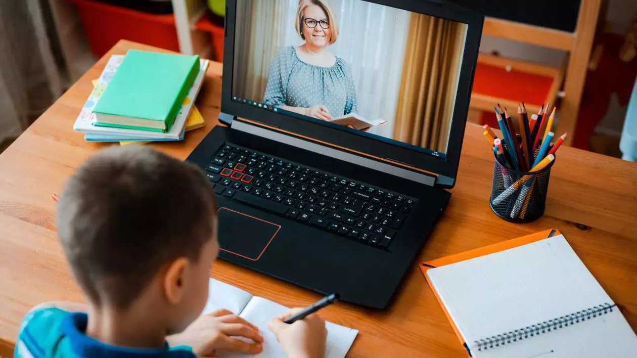 Un elev care stă cu spatele scrie pe caiet, iar în fața lui este un laptop deschis pe care comunică cu profesoara, care se vede pe ecran