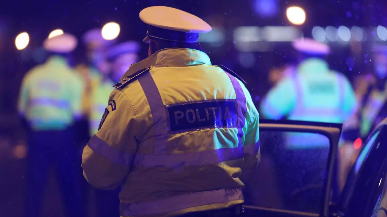 Polițist îmbrăcat în uniformă, fotografiat din spate, noaptea, lângă o mașină de Poliție cu ușa deschisă
