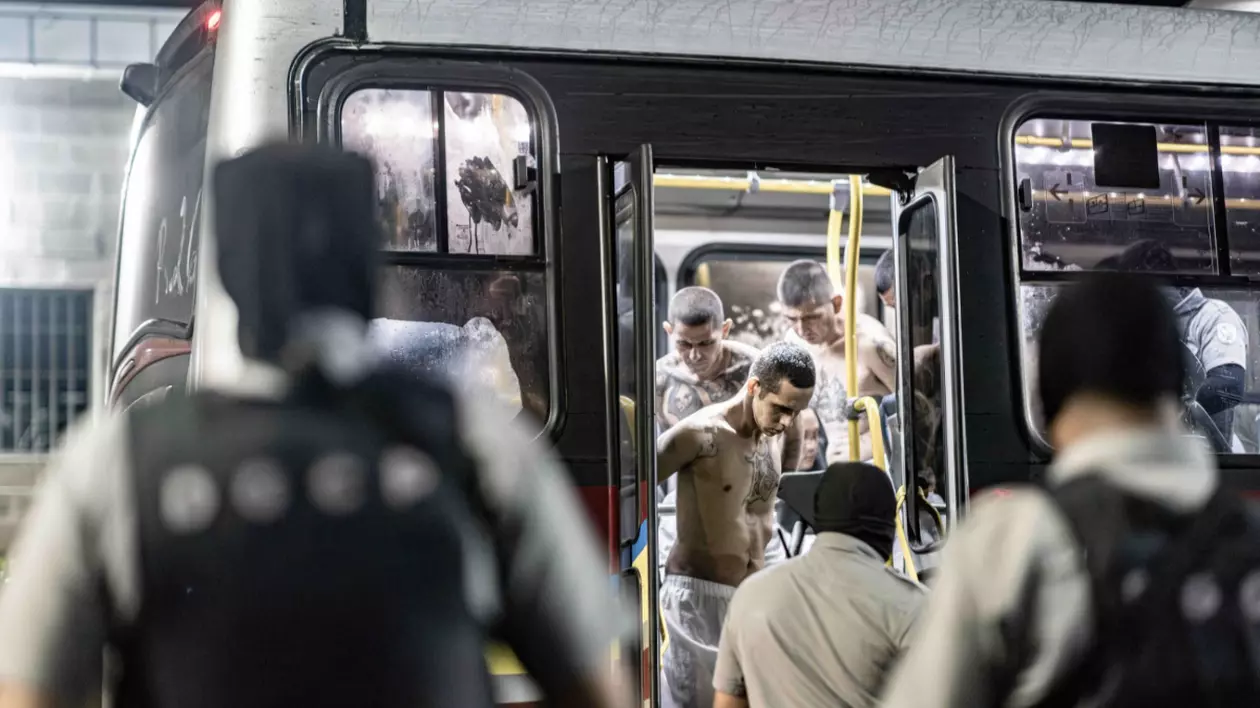 Grup de prizonieri cu bustul gol, mulți dintre ei având tatuaje vizibile, coborând dintr-un autobuz sub supravegherea unor agenți de securitate mascați.