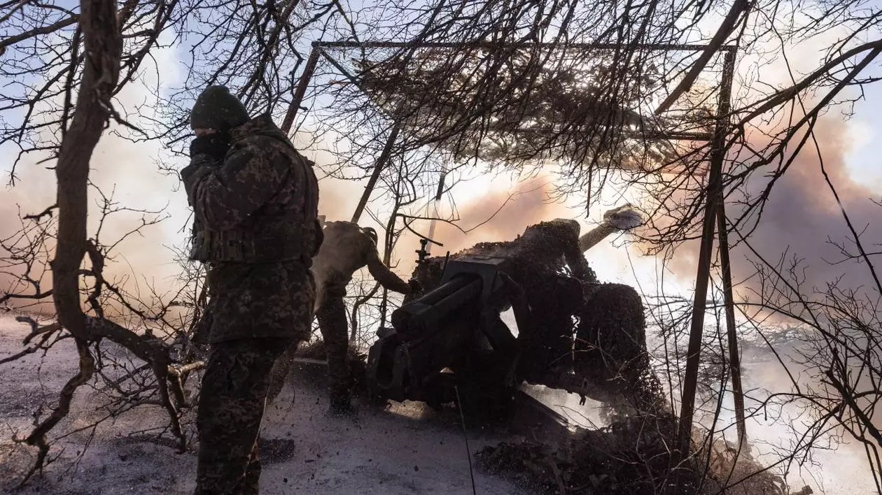soldați, îmbrăcați în uniforme de camuflaj, operează un tun de artilerie ascuns sub o plasă de camuflaj printre crengi