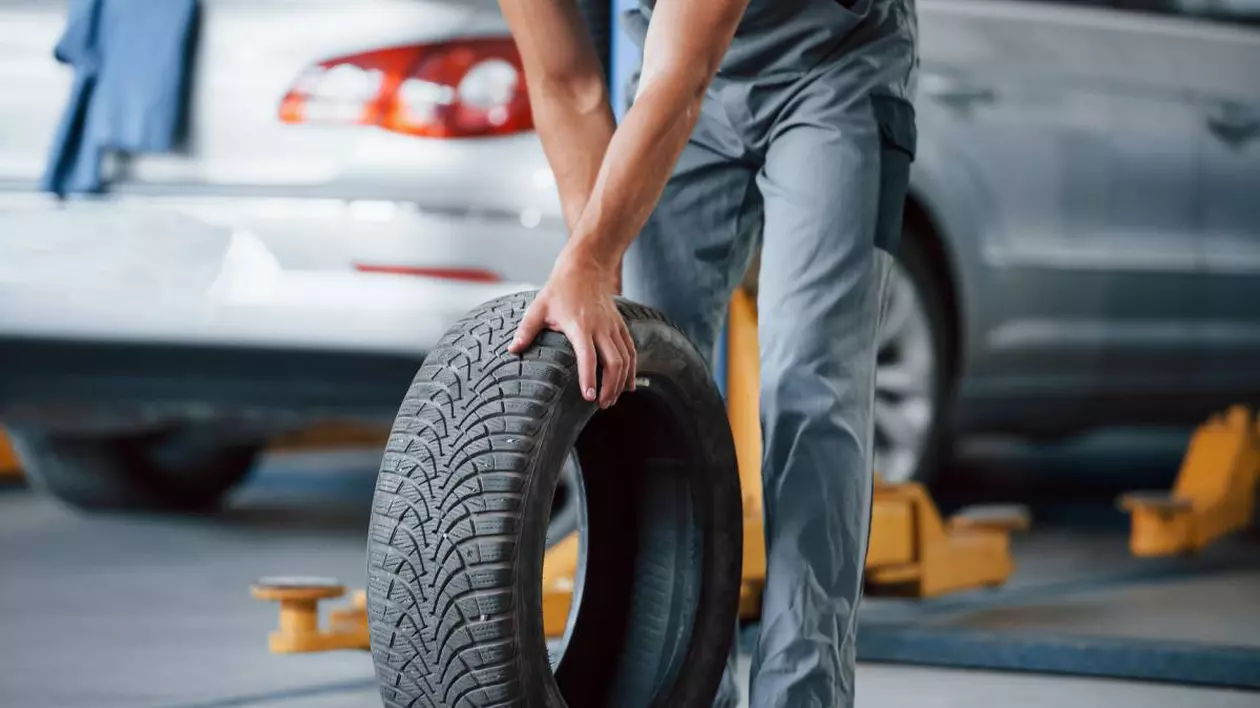 mecanic auto cu uniforma gri, invarte un cauciuc de masina. in spatele lui este o masina gri