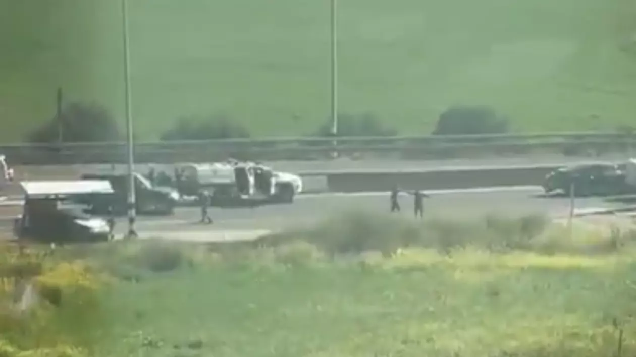Imaginea surprinde o scenă desfășurată pe o șosea, unde mai mulți oameni par implicați într-un incident. În prim-plan, se vede o stație de autobuz și mai multe vehicule oprite, inclusiv o camionetă albă cu ușile deschise.