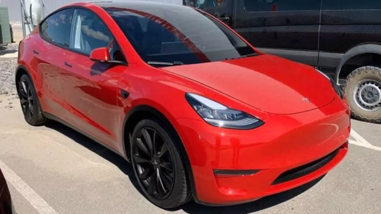 O mașină Tesla roșie, într-o parcare, alături de un microbuz negru