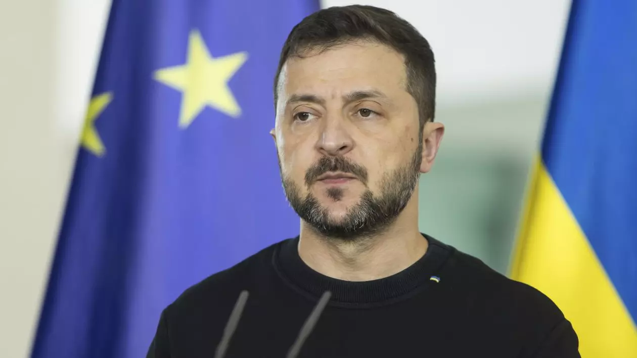 Volodimir Zelenski, președintele Ucrainei, într-o conferință de presă, îmbrăcat cu un pulover de culoare neagră, iar pe fundal steagurile Ucrainei și Uniunii Europene