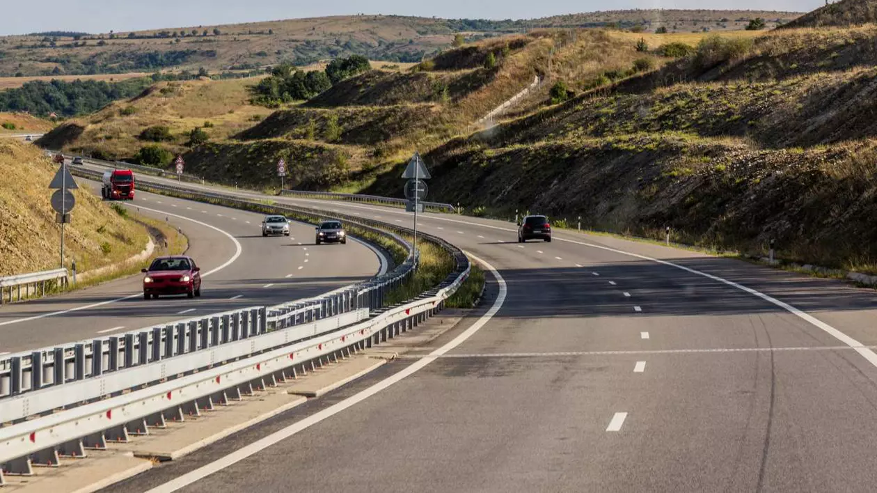 Autostradă din Bulgaria, printre dealuri, cu doua benzi pe sens, cu câteva autoturisme și un camion. Pe marginea drumului se observă parapete de siguranță