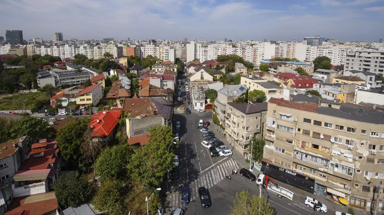 O intersecție din București, mai multe mașini, o trecere de pietoni, blocuri, un spațiu verde