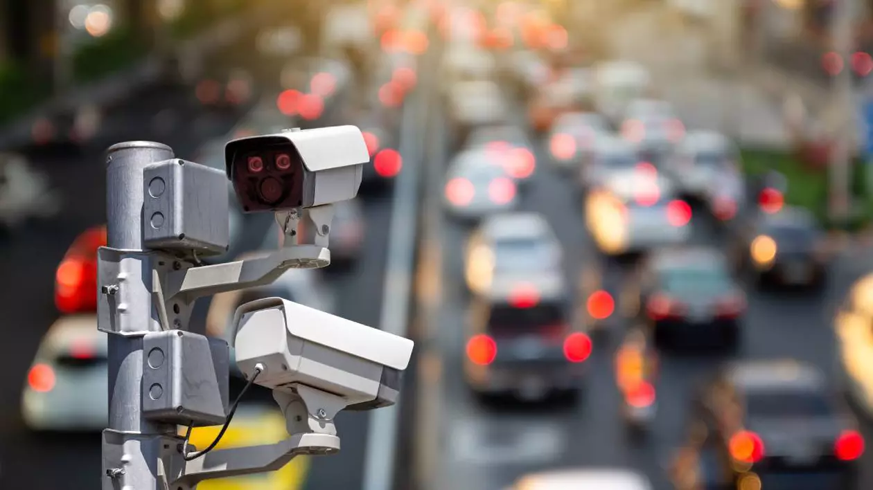 Trei camere de supraveghere montate pe un stâlp metalic, îndreptate către o șosea intens circulată. Fundalul este ușor blurat, dar se observă un trafic aglomerat, cu mașini aflate în coloană și luminile roșii ale stopurilor aprinse.