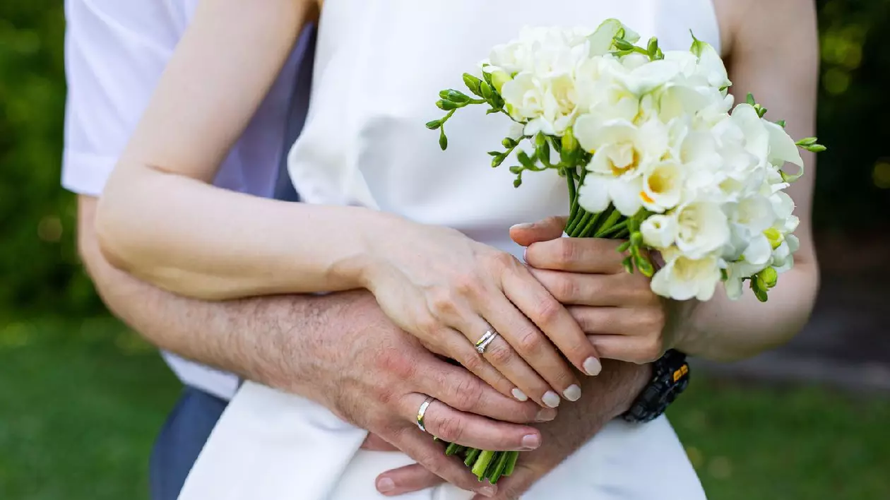 O mireasă și un mire se țin de mână și au un buchet de flori albe. Mireasa poartă o rochie albă, iar mirele poartă un costum. Scena este veselă și romantică, deoarece cuplul se îmbrățișează