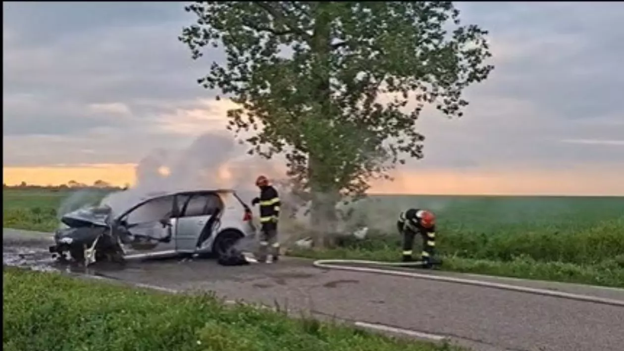 mașină în flăcări din care iese fum gros, negru, înconjurată de pompieri care încearcă să stingă incendiul