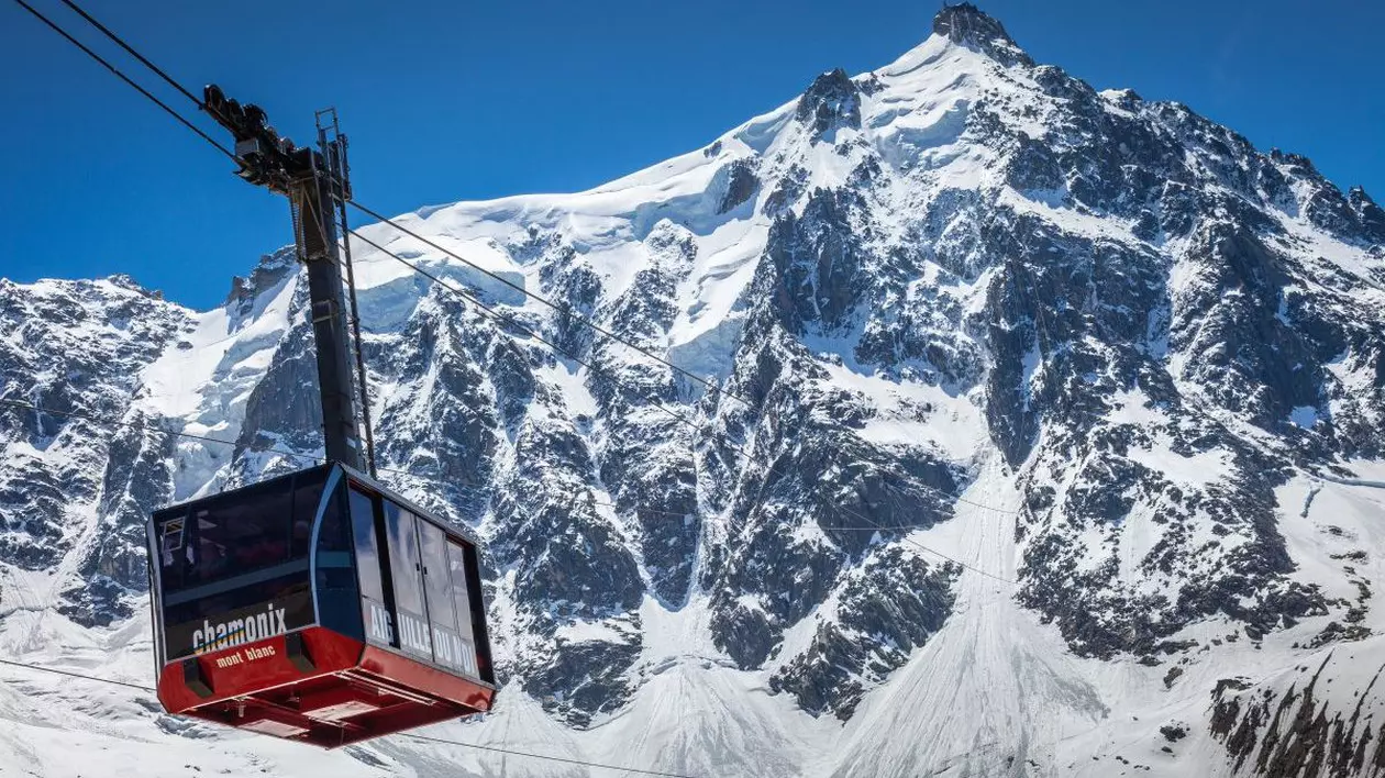 O telecabină negru cu roșu este pe fir către Aiguille du Midi, Masivul Mont Blanc, Chamonix, estul Franței, iar în depărtare se văd munții acoperiți cu zăpadă
