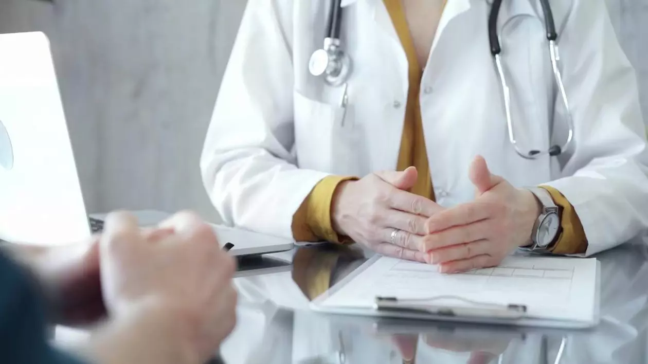 Medic îmbrăcat în halat alb, purtând un stetoscop în jurul gâtului și un ceas de mână, gesticulând ușor deasupra unei mese pe care se află o fișă medicală și un laptop deschis.