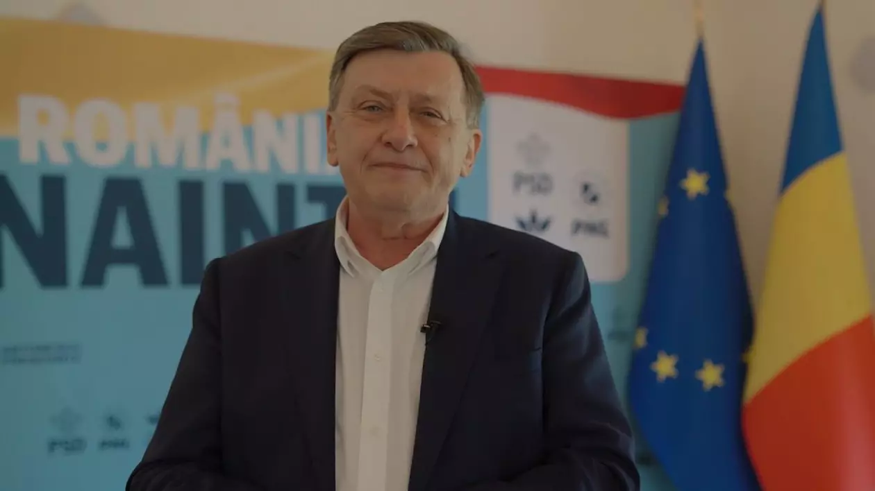 Crin Antonescu, în cămașă albă și sacou desfăcut, în timp ce zâmbește, cu steagurile României și Uniunii Europene în fundal